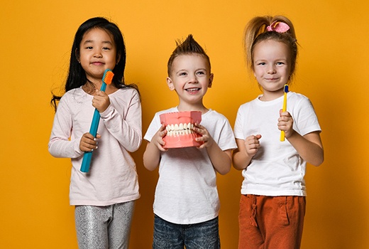 Three children holding oversized toothbrushes and model of teeth in front of goldenrod background